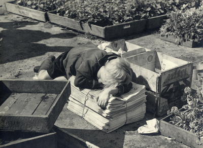 98205 Afbeelding van een jongetje, slapend op een stapeltje kranten, in een kwekerij op een onbekende locatie, mogelijk ...